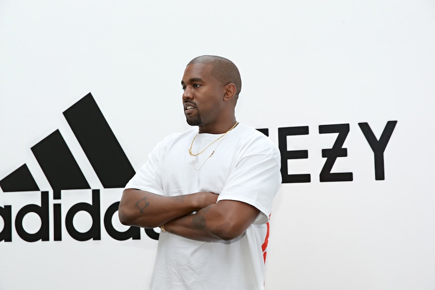 Ye wearing white and standing with arms crossed in front of an adidas and Yeezy sign
