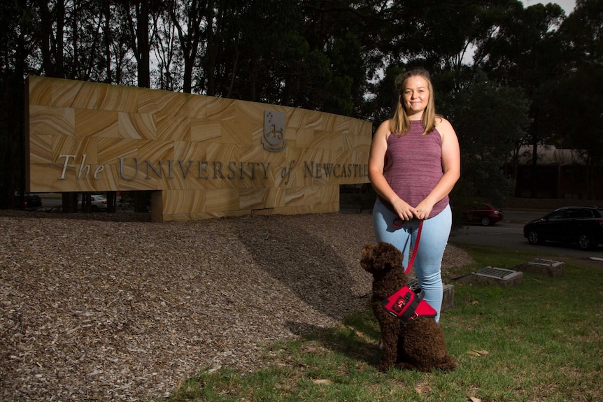 A young woman standing with a dog in front of the University of Newcastle sign