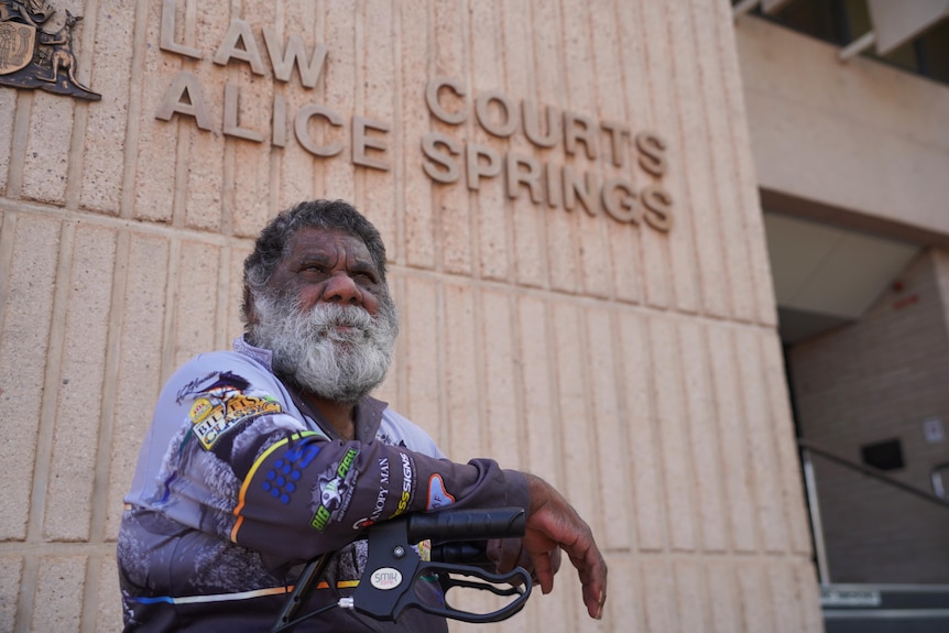 A man sits in a walker in from of the Alice Springs law court. His expression is serious.