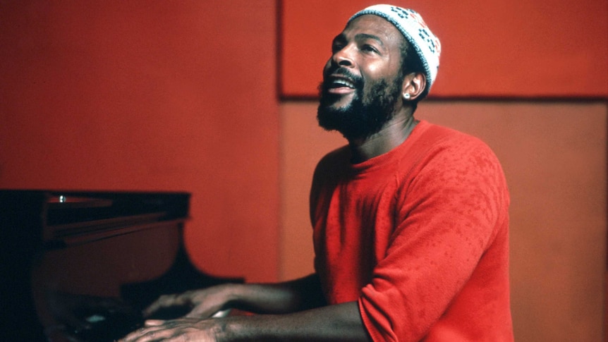 Marvin Gaye wears a red tracksuits and sits down at a piano in a red studio. He is wearing a white beanie and singing.