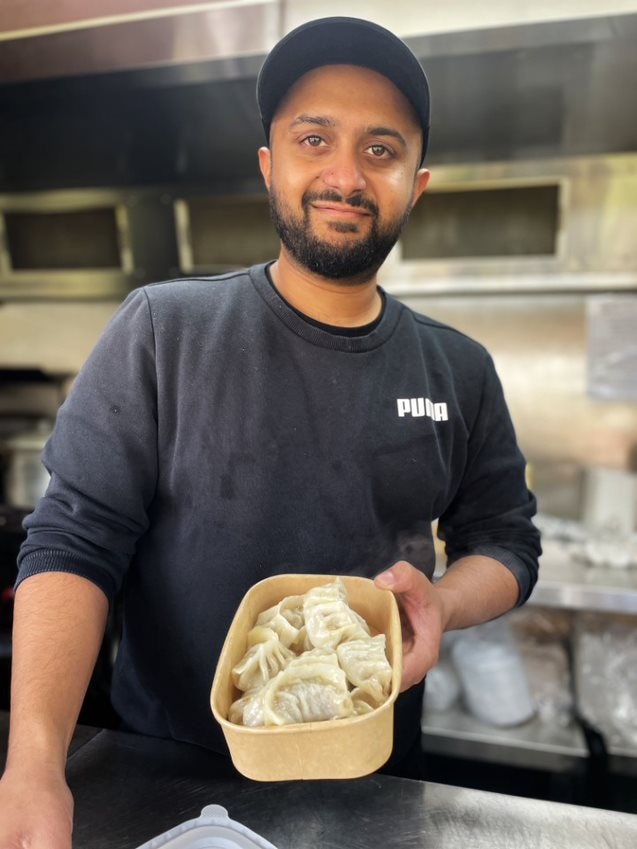 A man in a black cap smiles and holds out a container of Nepali momo.