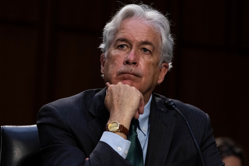 Man in a suit leans forward in his chair resting his chin on his fist. 