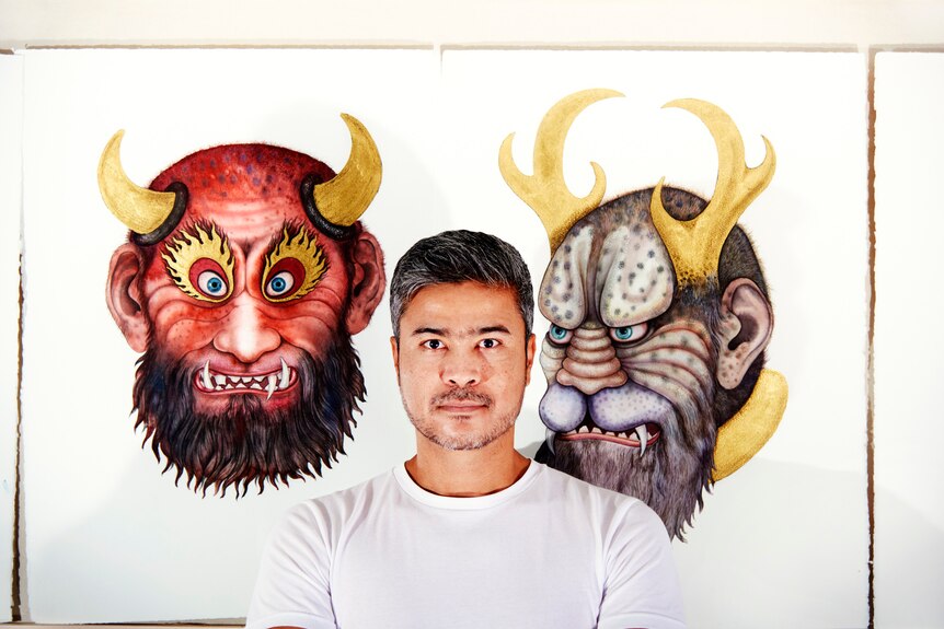 Artist Khadim Ali standing in front of paintings of two fantastical characters, his head framed by devil horns