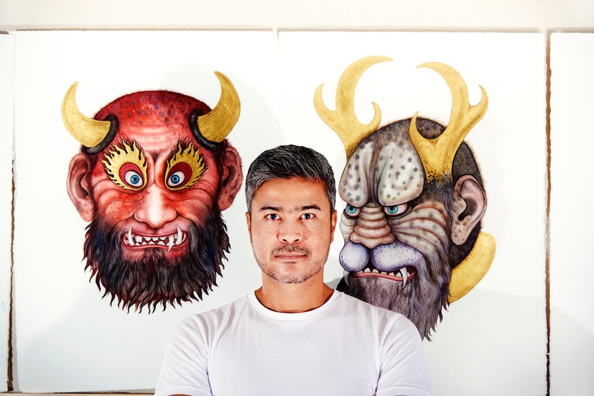 Artist Khadim Ali standing in front of paintings of two fantastical characters, his head framed by devil horns