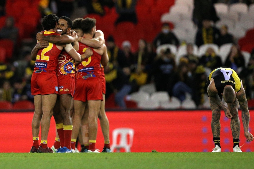 Four Gold Coast Suns AFL players embrace as a Richmond player stands in the background.
