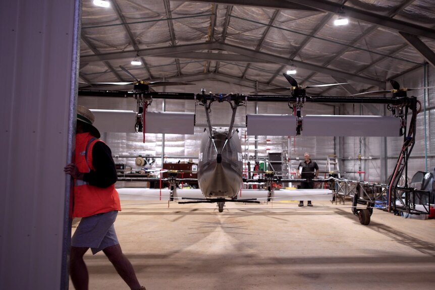 A person in a fluro jacket pushes open the door of an aircraft hangar to reveal a small electric aircraft