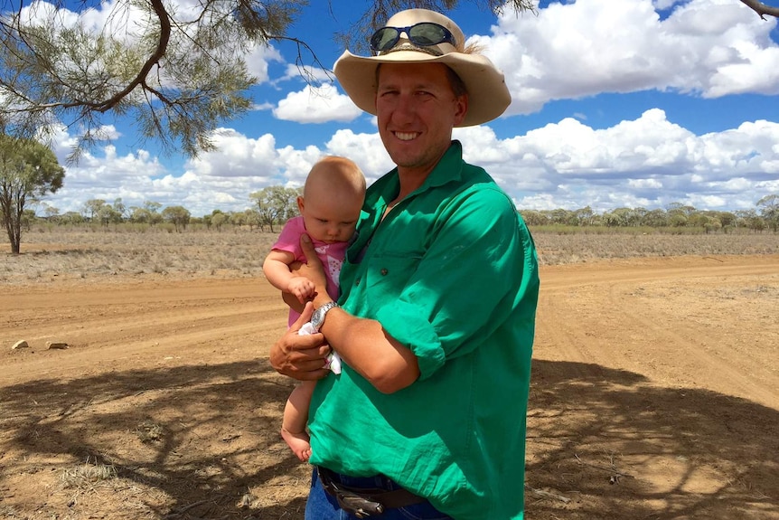 A young farmer holding his baby daughter stands under the shade of a tree in a dry paddock.