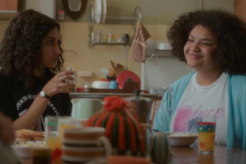 Ambika and Amber sit at a kitchen table in the morning, Ambika looking to Amber who smiles at someone off screen.