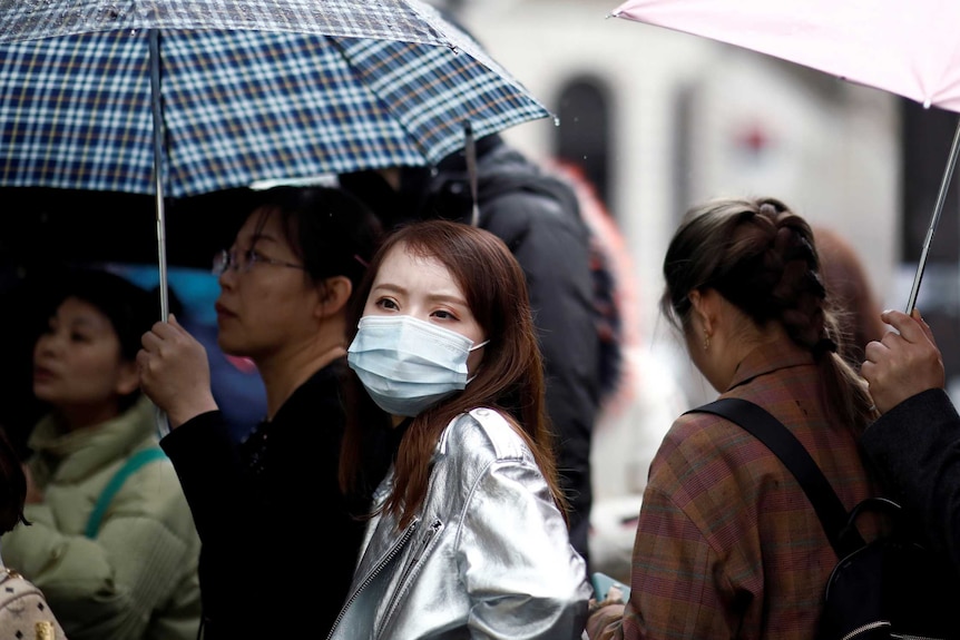 a woman wears a mask in a crowd holding umbrellas