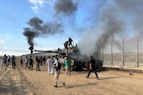 People stand as smoke billows from a tank by a large fence