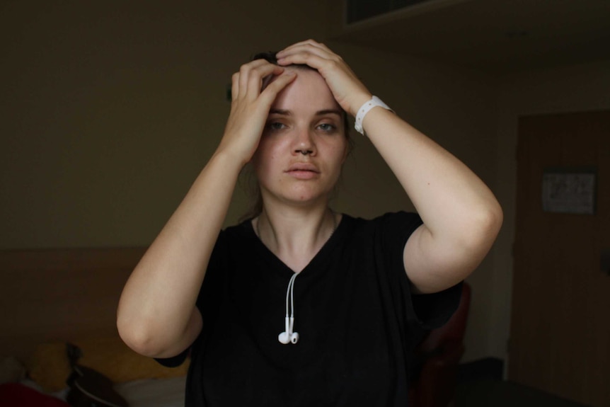 A young woman with brown hair wearing a black shirt with white headphones looking at the camera.