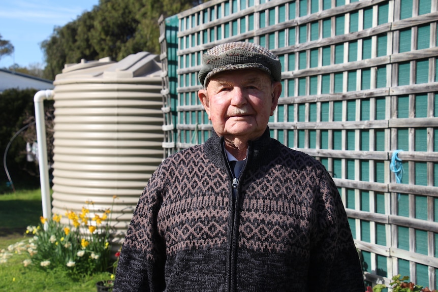 An older man in a cardigan and cap stares at the camera with a water tank and lattice behind him