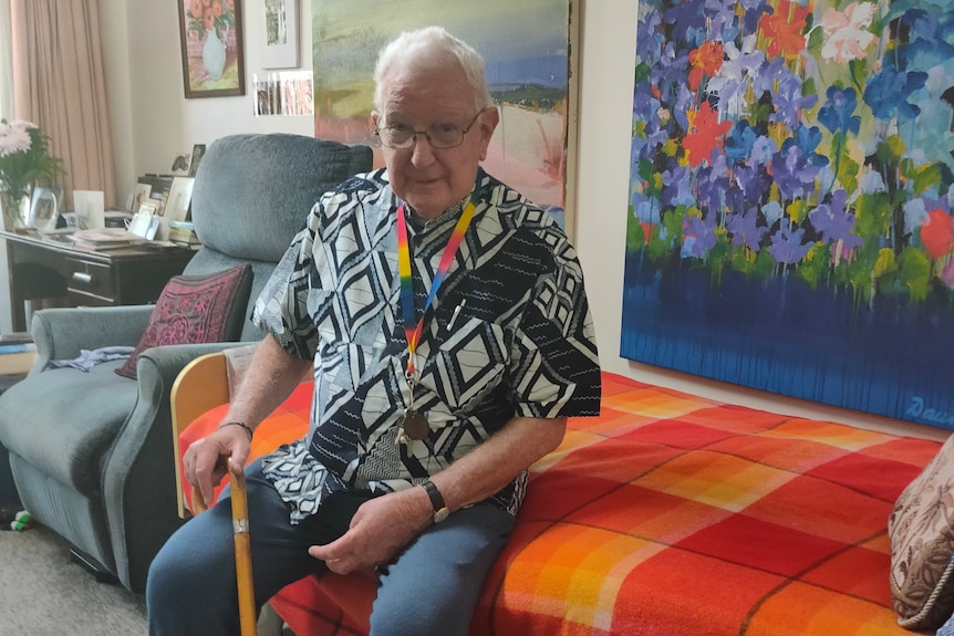 Don Maynard sitting on a bed holding a cane with paintings on the wall behind him.