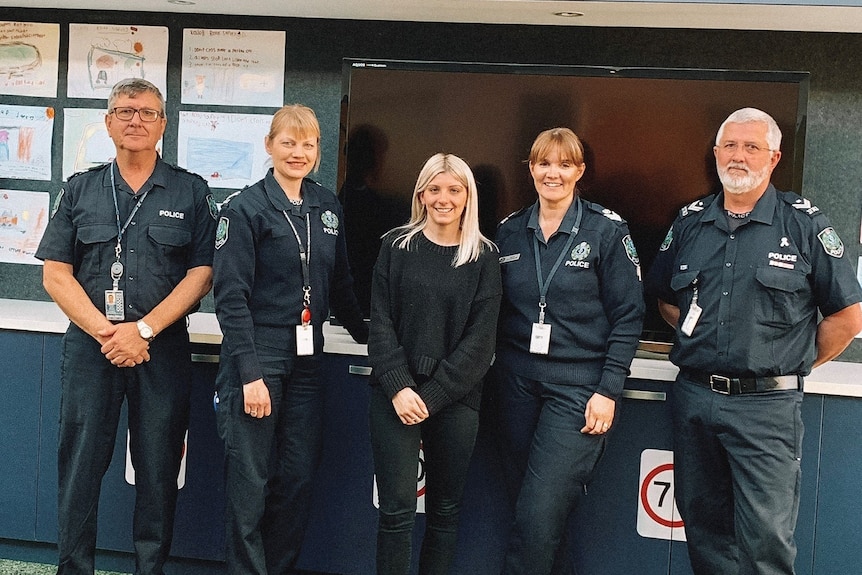 A group of police in uniform stand next to a woman in a black jumper in an office. 