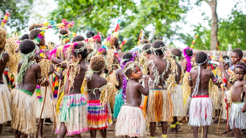 A group of children in grass skirts with one looking back at the camera. All have bare feet and stand on bare earth.