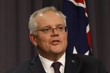 A man with grey hair and glasses wearing a suit and tie speaking at a lecturn in front of an Australian flag