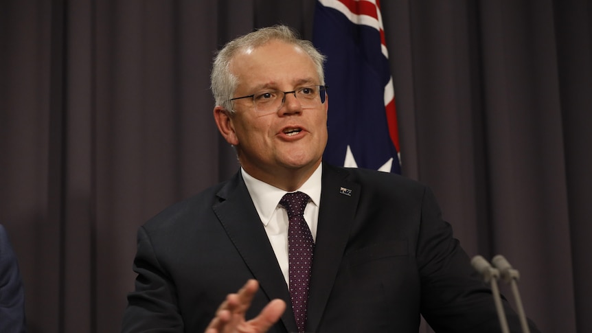 Un uomo con i capelli grigi e gli occhiali in giacca e cravatta parla davanti a una conferenza davanti a una bandiera australiana