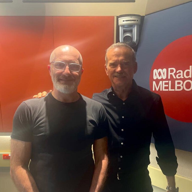 Two smiling men stand in front of the ABC Melbourne logo