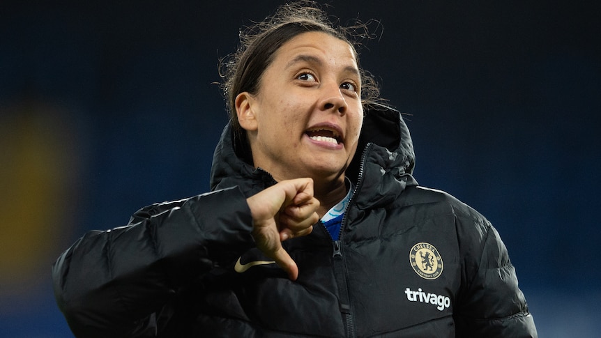 Sam Kerr points at herself with her thumb while wearing a big puffy jacket over her Chelsea jersey.