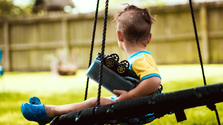 A small child sits on a swing in a backyard with his head turned away from the camera