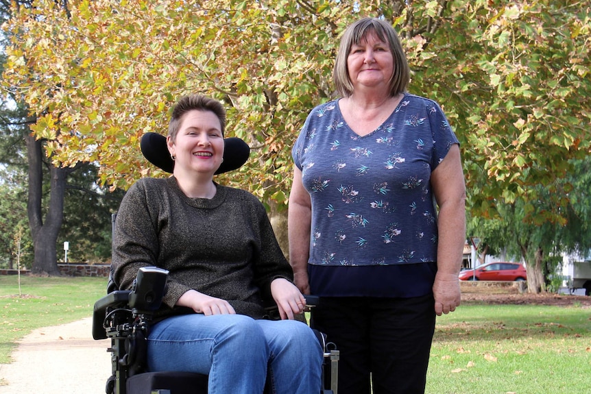 Anj Barker (left) sitting in a park in her wheelchair with her mother Helen standing beside her.