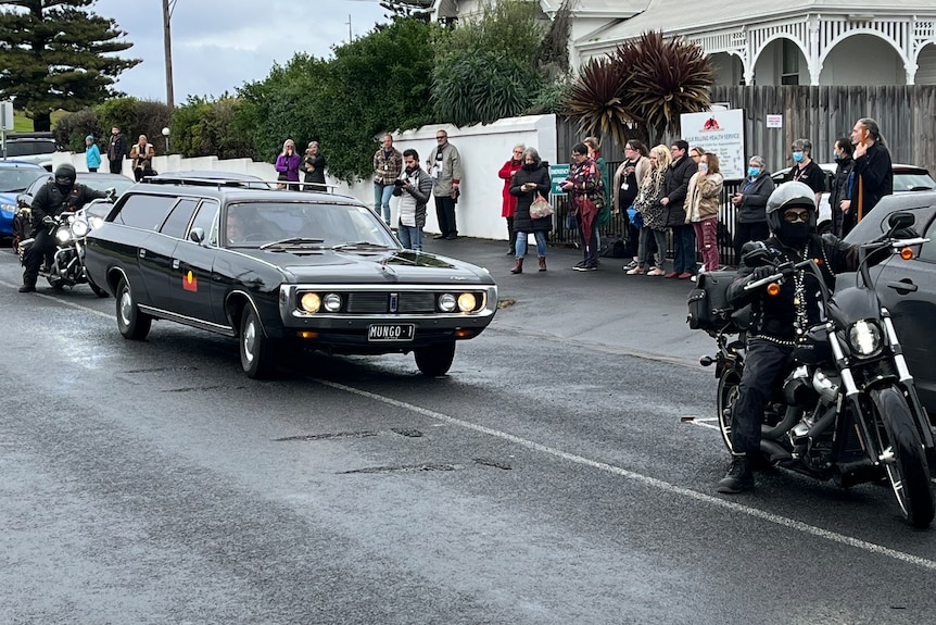 Motorbikes and black hearse on the road, people line the footpath.