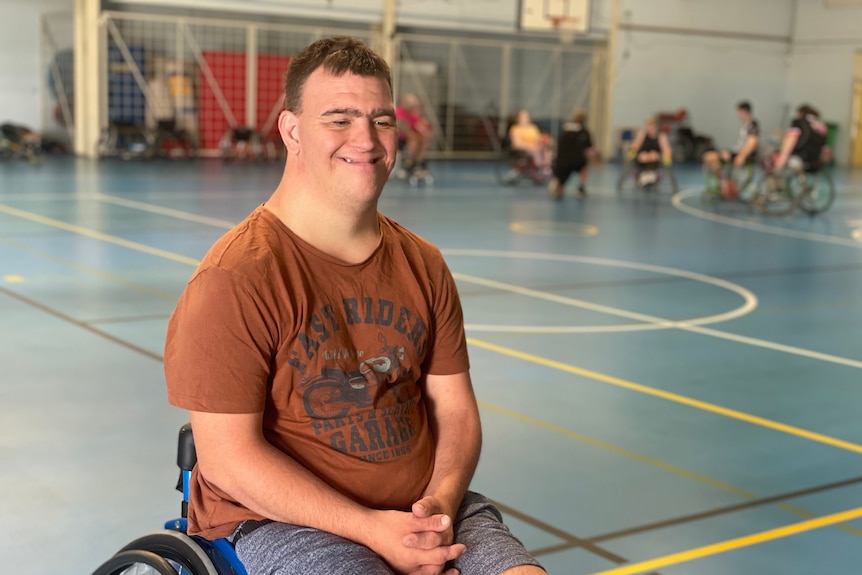 A young man smiles while a game of wheelchair basketball is played behind him