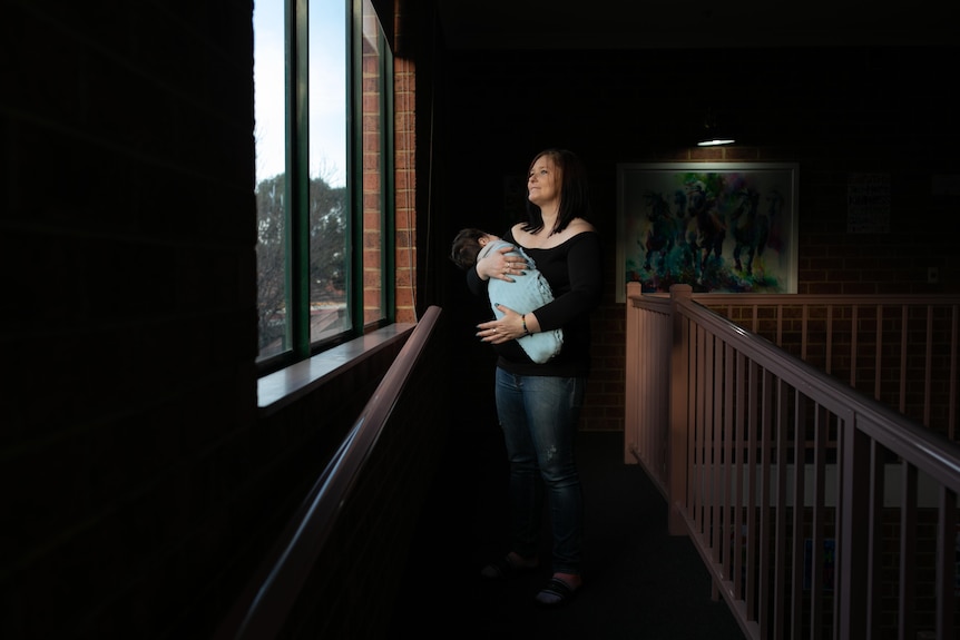 A woman stands, holding a baby in her hands, looking out a window. The room around her is dark.