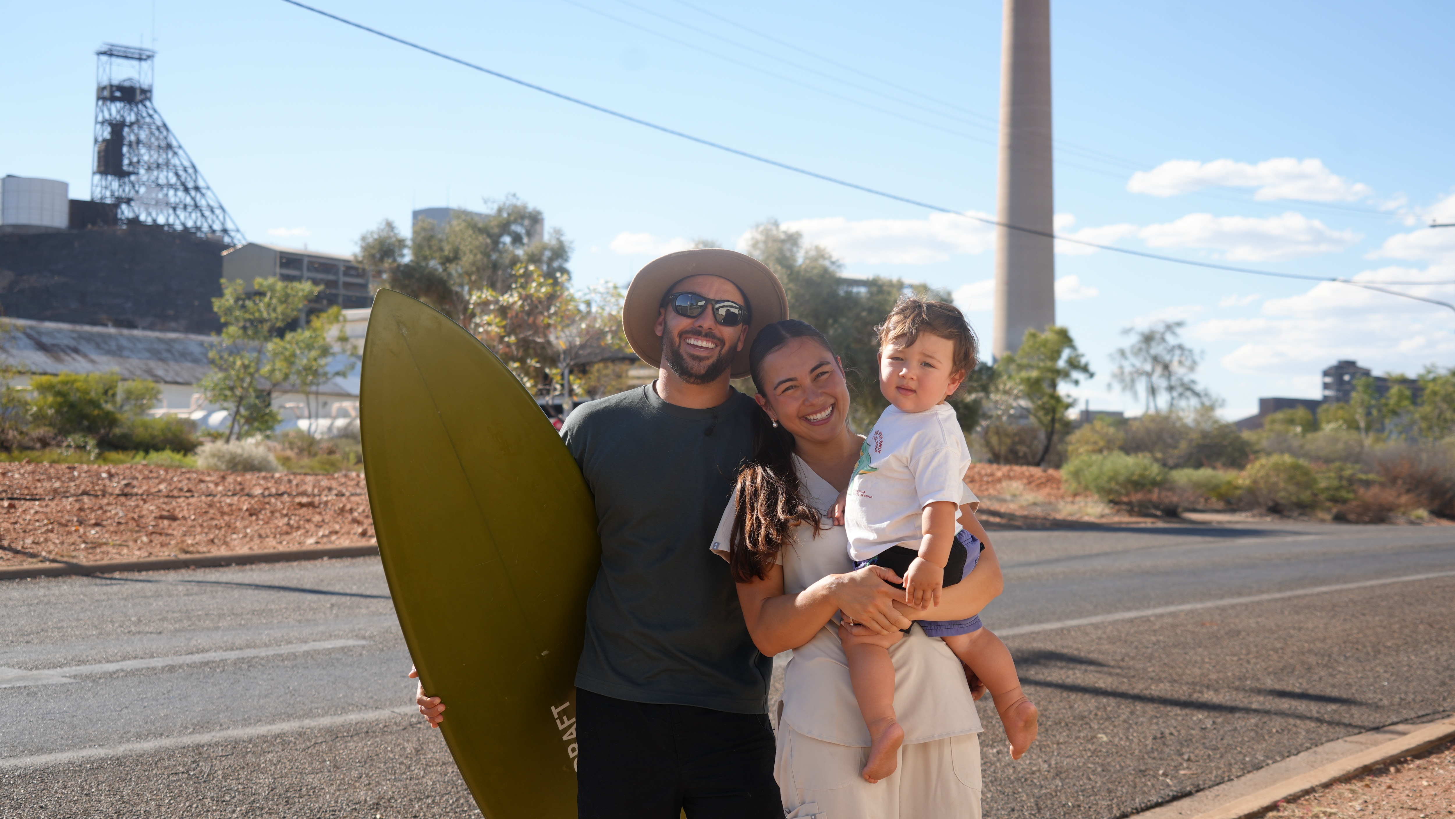 A man wearing a hat and sunglasses holds a surfboard as he stands alongside a woman carrying a child