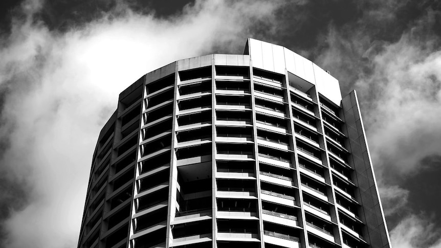 A black and white photo shows the curvature of  Harry Seidler's Shell House tower set against fast-moving clouds.