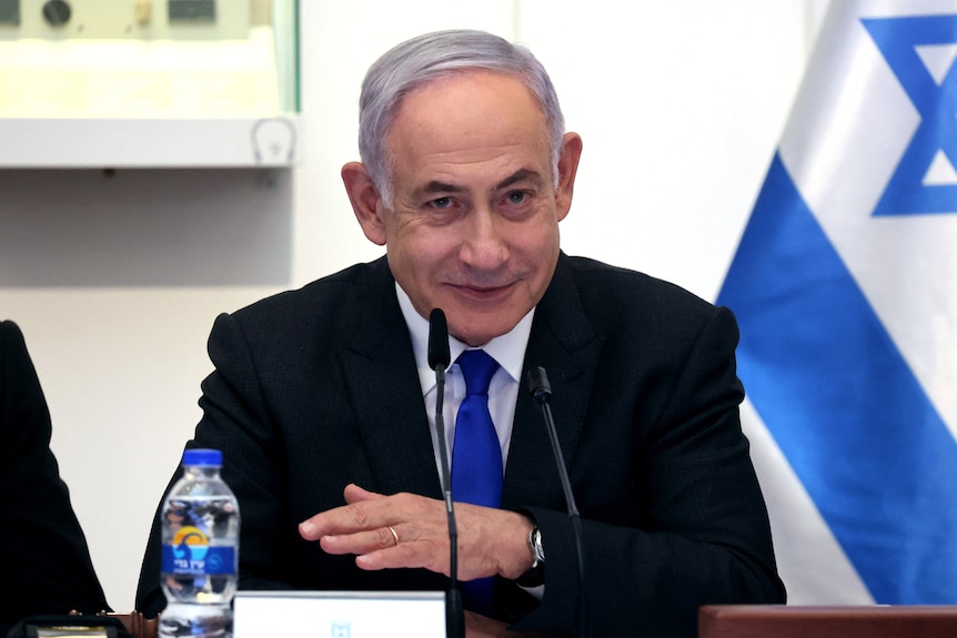 Benjamin Netanyahu grins as he sits behind two small microphones mounted to a desk in front of an Israeli flag.