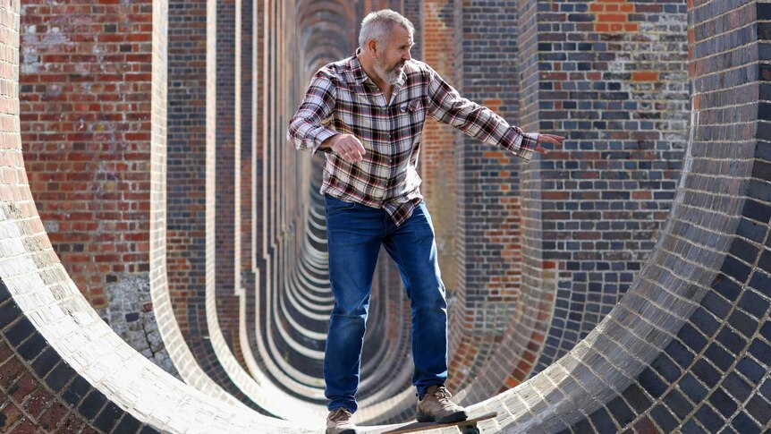An older man skateboarding underneath Ouse Valley viaduct. He is wearing a flannel shirt and is holding out his hands to balance