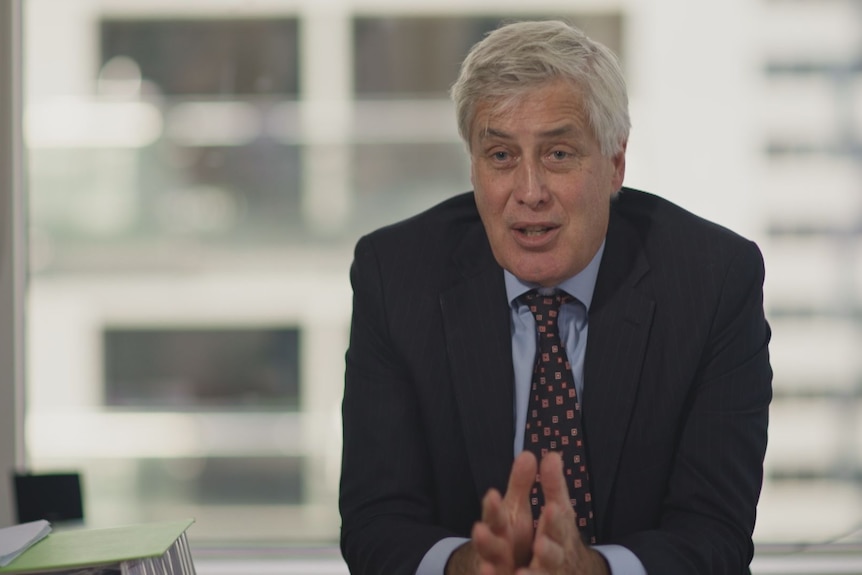 John Ribbands in a suit and tie sitting at at desk with his hands close together mid-sentence. The background is out of focus