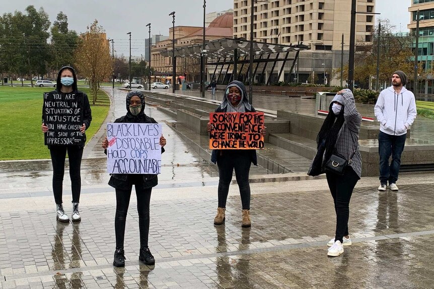 Five people stand space apart in a square in the rain, some holding anti-racism signs.