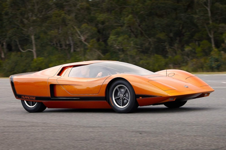 An orange Holden Hurricane concept car, with a man driving it, on a road with a background of trees and shubs.