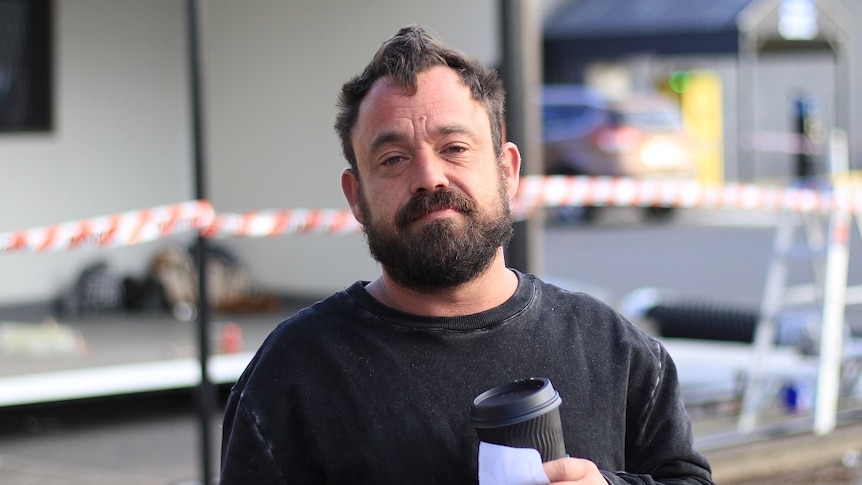 A man holds a takeaway coffee cup in front of a demountable building.