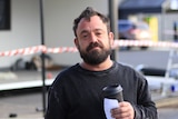 A man holds a takeaway coffee cup in front of a demountable building.