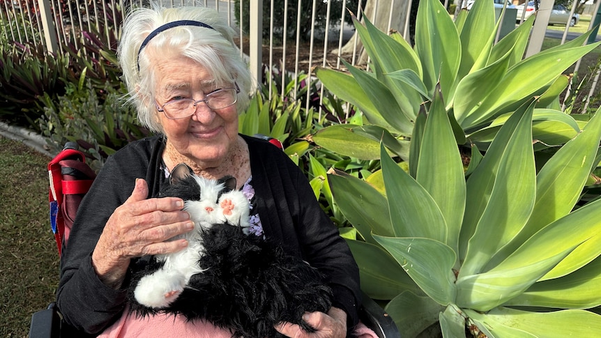 An older woman with short white hair smiling at the camera, holding a robotic cat on her lap