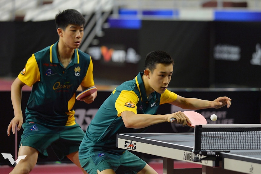 Two table tennis players play in a doubles match, the player in front prepares to hit the ball with his paddle