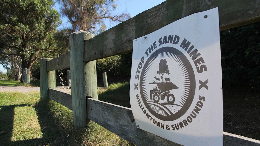 A sign that says "stop the sand mines" is posted to a wooden fence with trees in the background