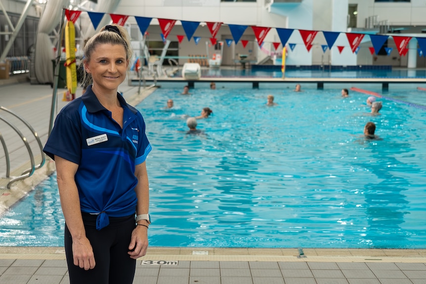 Woman smiling at camera wears a blue polo shirt and stands to the left of an aqua aerobics class taking place.