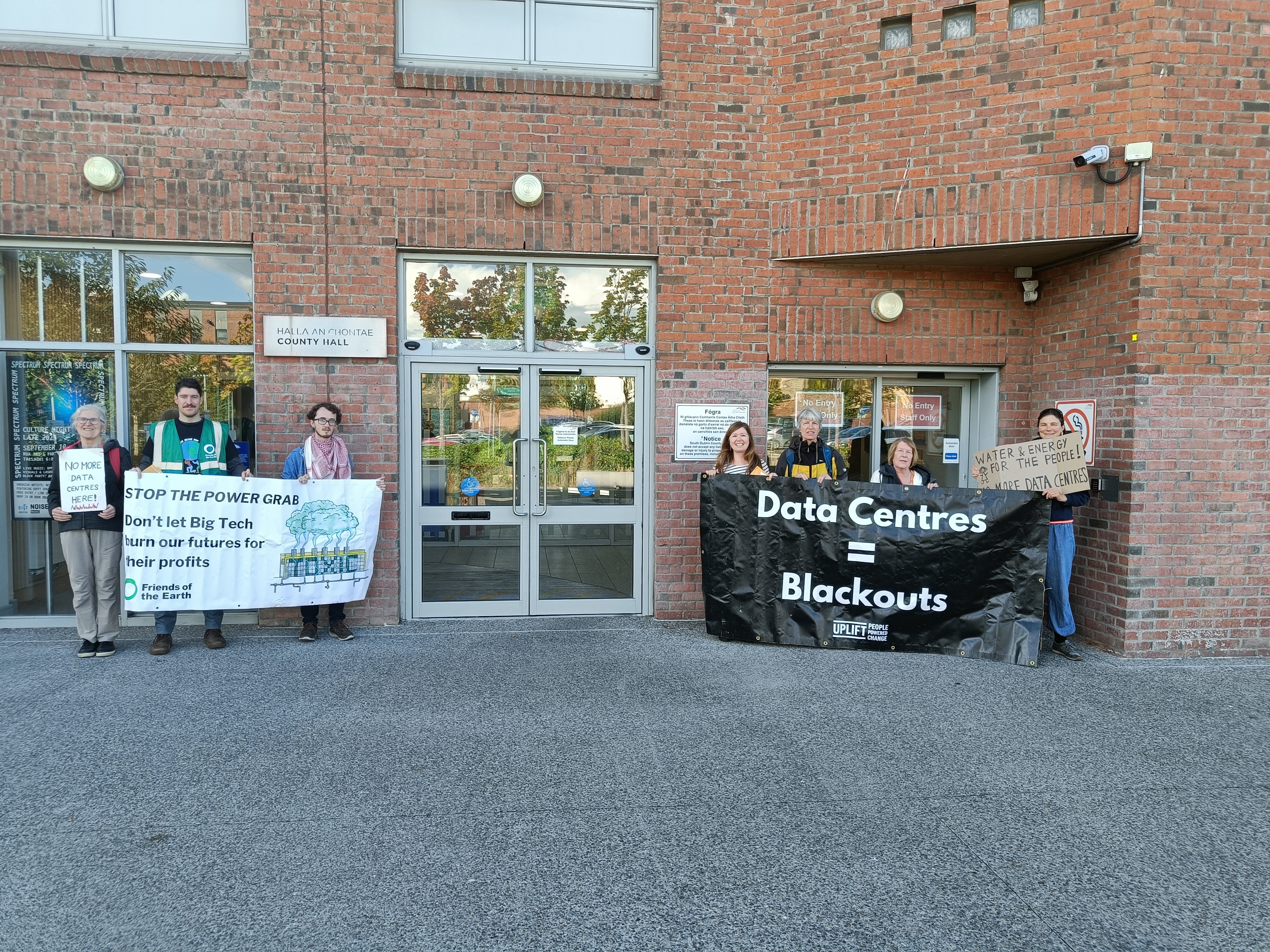 A small cluster of people outside a building hold banners opposing data centres. 