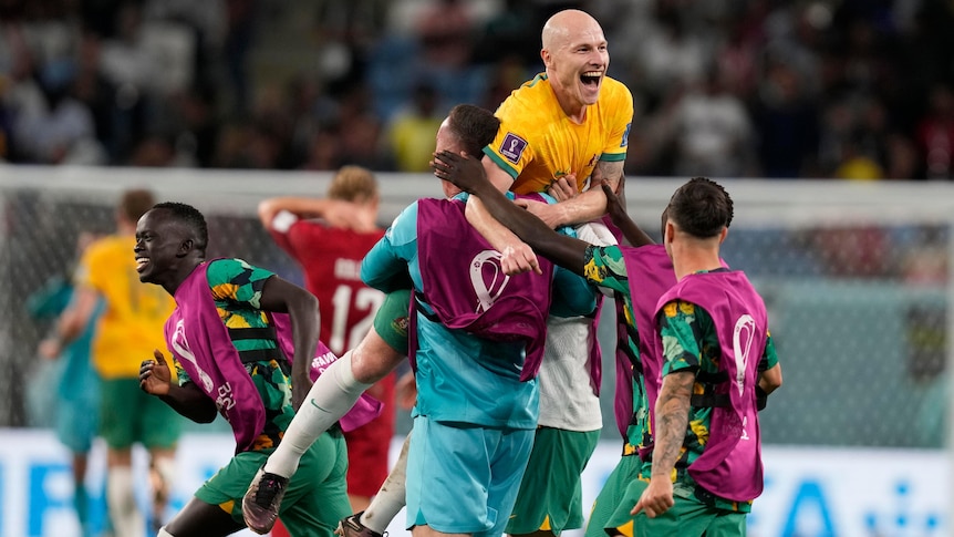 Aaron Mooy smiles and jumps in the air, held by a teammate as others run around him