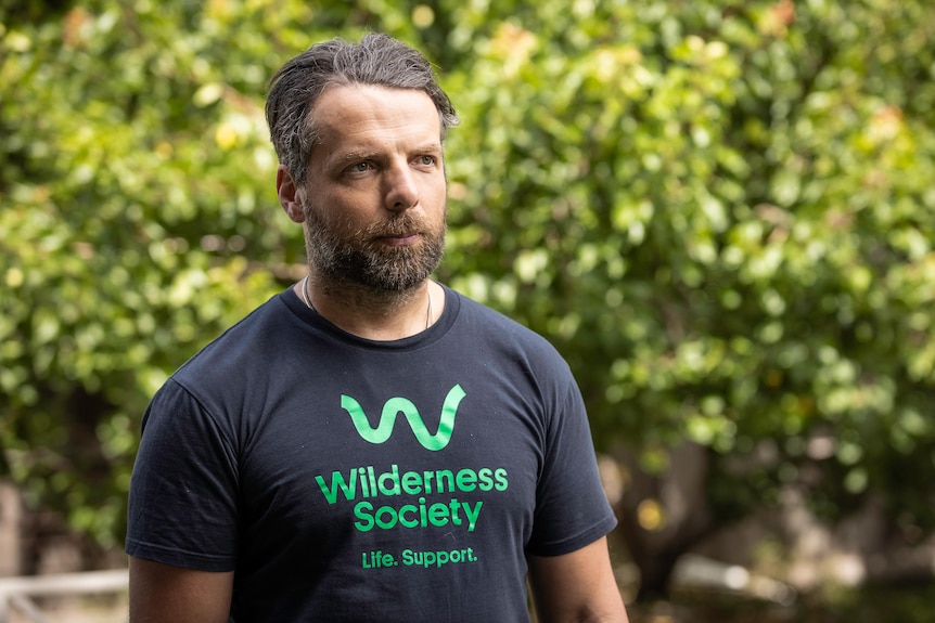 Un hombre con una camiseta negra que dice Wilderness Society Life.  Puestos de apoyo frente a la vegetación desenfocada