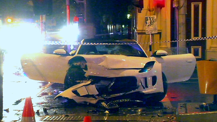 A white sports car sits on the road with its front smashed in and doors flung open after a crash.