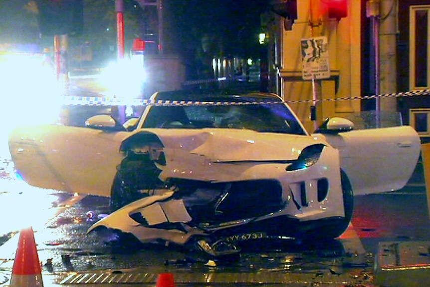 A white sports car sits on the road with its front smashed in and doors flung open after a crash.