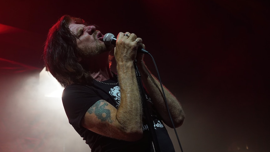 Tex Perkins stands on stage, holding tightly onto a microphone with his eyes closed. He's wearing a black t-shirt.