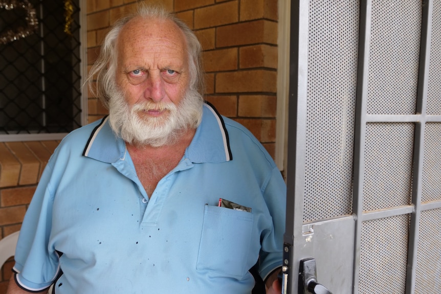 A man in a blue polo shirt with grey hair and a beard, standing next to a door.