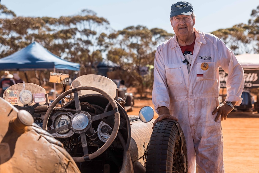 A man wearing dirty overalls standing next to a classic car.  