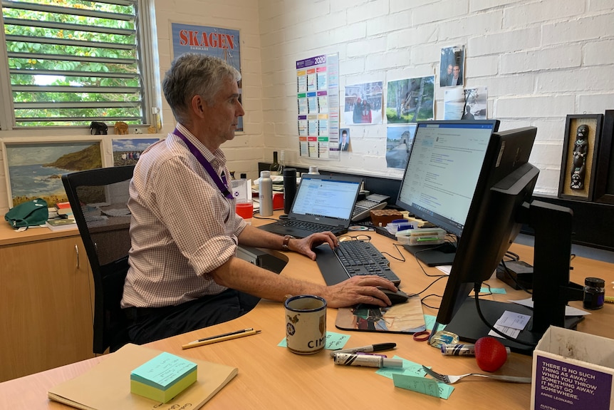 A man sits at a desk with two computer screens sits.